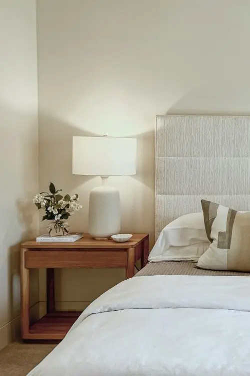 helles Polsterbett mit Nachttisch aus Holz und eingeschalteter, weißer Tischleuchte